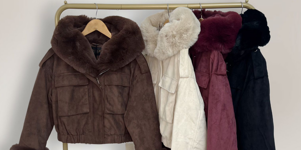 Wholesale women’s suede faux fur hooded bomber jackets in brown, cream, burgundy, and black cropped winter outerwear with detachable fur trim from AW25 wholesale collection for retail buyers. Jackets hung on a rail.