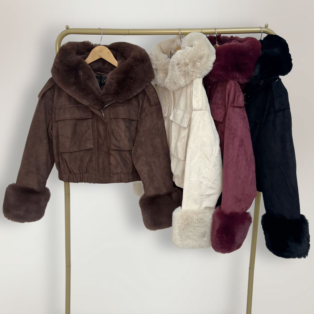 Wholesale women’s suede faux fur hooded bomber jackets in brown, cream, burgundy, and black cropped winter outerwear with detachable fur trim from AW25 wholesale collection for retail buyers. Jackets hung on a rail.